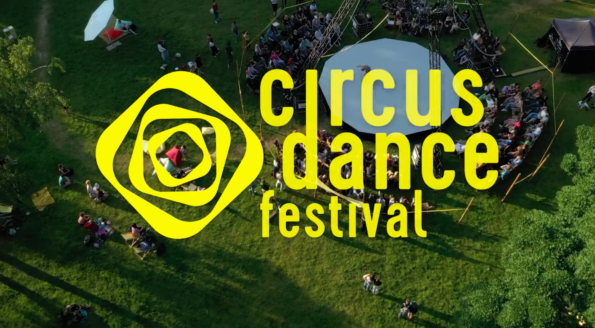 Luftaufnahme eines grünen Geländes mit einem Zirkuszelt und Menschenansammlung beim "Circus Dance Festival" in großen gelben Buchstaben darüber.