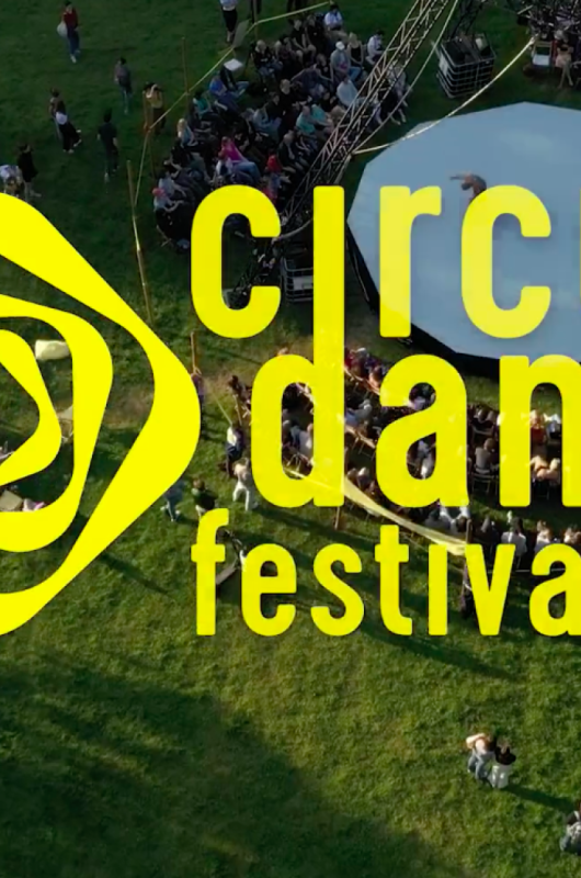 Luftaufnahme eines Zirkus-Tanzfestivals auf einer Wiese mit einem Bühnenbereich und Logo.