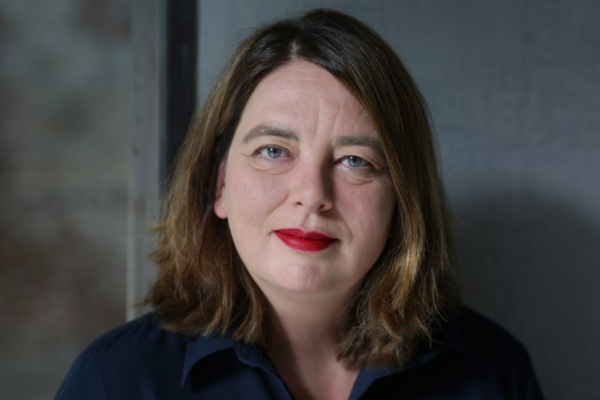 Eine Frau mit schulterlangem braunem Haar und rotem Lippenstift blickt direkt in die Kamera.