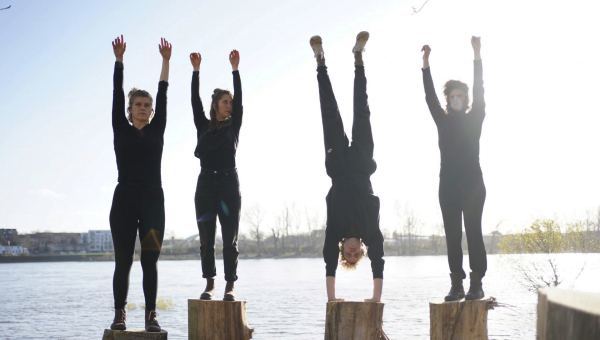 Vier Personen stehen auf Baumstümpfen am Wasser, eine davon macht einen Handstand.
