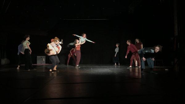 Eine Gruppe von Darstellern tanzt auf einer Bühne in einem dunkel beleuchteten Raum.