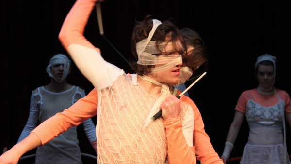 Eine Person in einem weißen und orangefarbenen Kostüm mit verbundenem Kopf hält einen Stock, während andere im Hintergrund stehen.