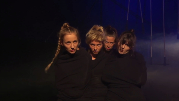 Vier Frauen stehen dicht zusammen in schwarzer Kleidung vor einem dunklen Hintergrund.