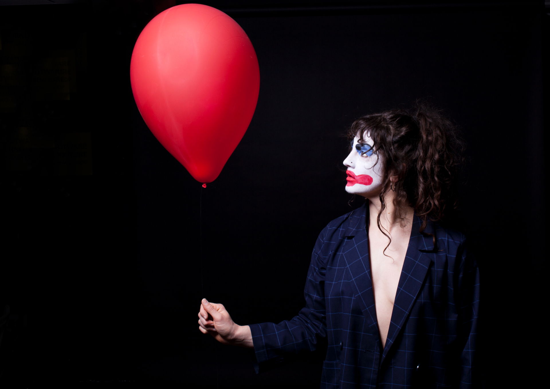 Eine Person mit Clown-Make-up hält einen roten Ballon vor einem dunklen Hintergrund.